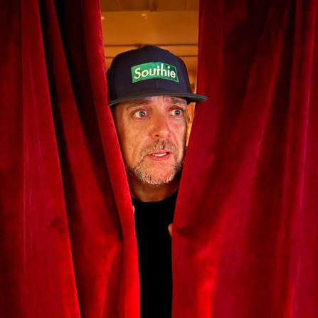 Gary Bolles peering through a red stage curtain, wearing a green Southie hat